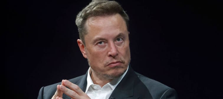 Elon Musk sits on stage in front of a black backdrop, looking perturbed.