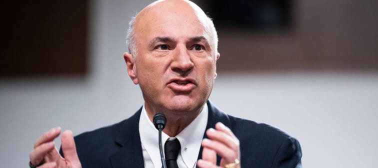 Kevin O'Leary wearing a suit, speaking into a microphone while gesturing his hands.