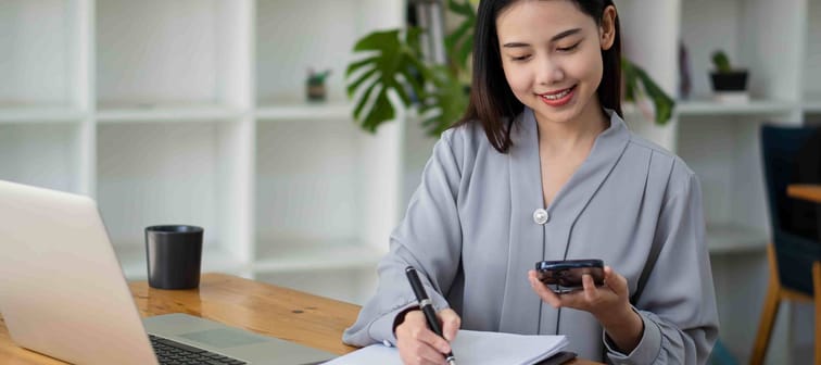 Asian businesswoman holding smartphone with charts or documents at work using business laptop calculator, financial accountant, money calculation, bank, loan, rent, payment.