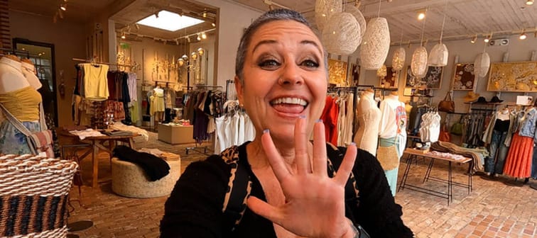 An older woman with short grey hair seen posing with a smile and her hand waving at camera in a clothing store.