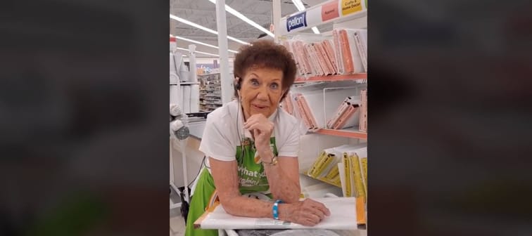 Older woman seen wearing Joann's Fabrics apron and leaning on table in aisle of the store.