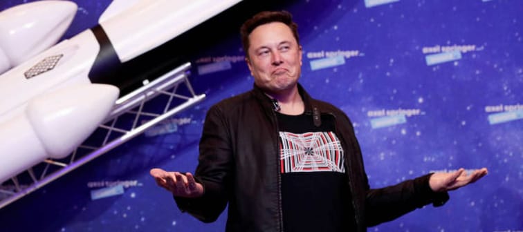 Elon Musk stands in front of a model rocket shrugging his shoulders.