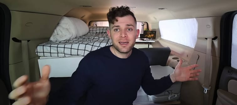 Young man seen posing with his arms outstretched in the back of a car, set up as a living space.
