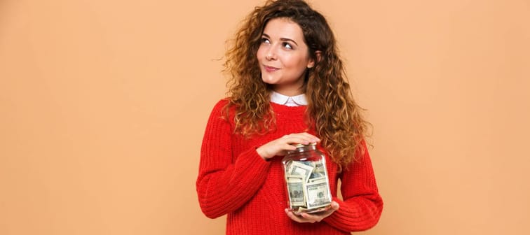 Portrait of a smiling young girl holding jar full of money banknotes and looking away isolated over beige background