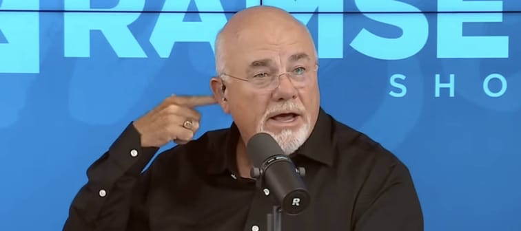 Dave Ramsey seen on set of his show, holding up a finger to his head, looking inquisitive.