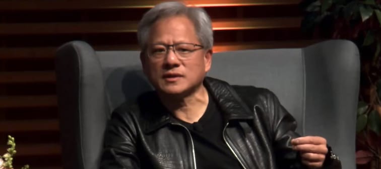 Jensen Huang seen in an armchair on a stage, speaking and gesturing with his hand.