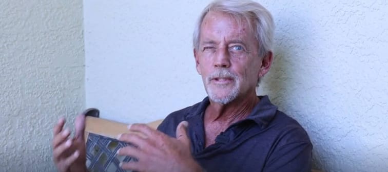 Older man sitting outside, gesturing and looking at the camera.