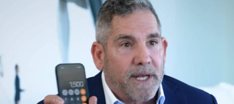 Grant Cardone sits in an office, holding up a calculator on his phone for the camera.