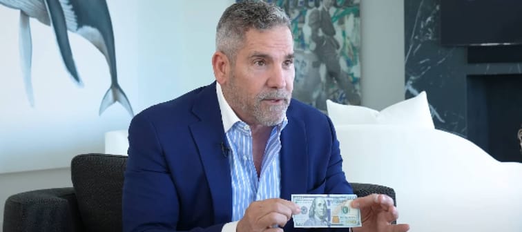 Grant Cardone seen sitting in a living room, holding up a $100 bill in his hands.