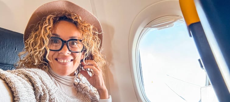 Woman with curly hair holds camera and smiles for selfie while sitting on airplane.