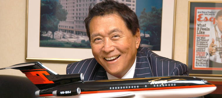 Robert Kiyosaki poses with a model airplane, smiling in an office.