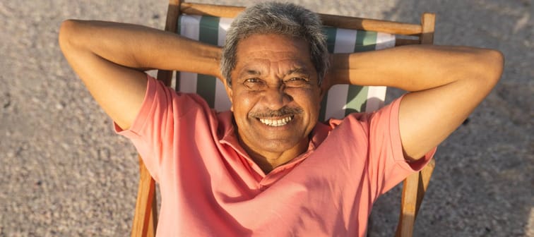 An older gentlemen smiles while sitting in a chair on the beach.