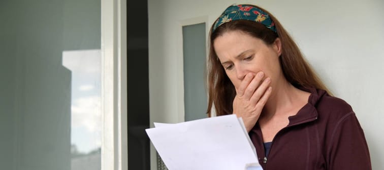 Shocked adult woman (female age 25-35) reading a letter outside a home front door.