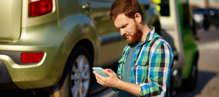 A man checks his phone while a tow truck drives away with his vehicle.