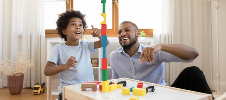 Black little son and happy dad building high toy tower together