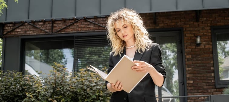 stylish blonde real estate agent looking at documents in folder near contemporary urban building