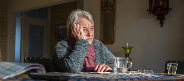 Older woman sits looking downcast with head in hand at dining room table.