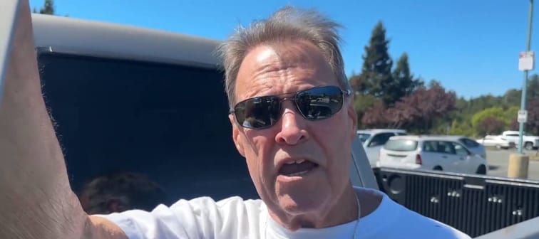 Man speaks to camera standing in front of truck, looking outraged.