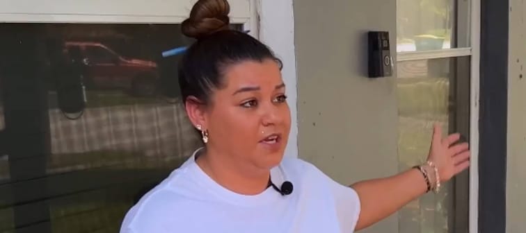 Woman in white t-shirt speaks to camera in front of house, gesturing and looking frustrated.