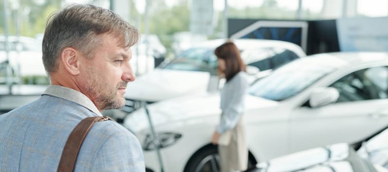 Contemporary mature man in smart casualwear keeping hand on front of new car of white color and looking at it while choosing vehicle