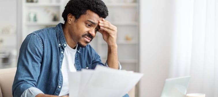 Man seated with papers in hand exhibits a worried expression while looking at his laptop at home