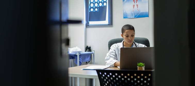 Doctor sitting at desk, wearing white coat and looking at computer.