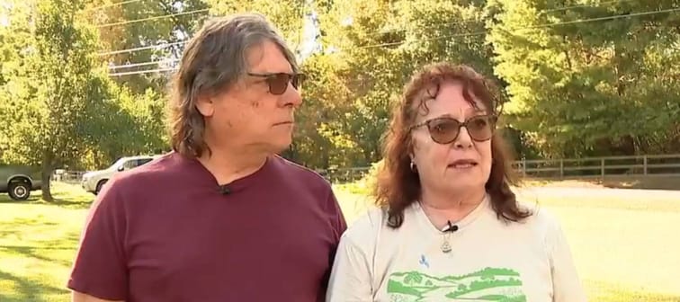 Man and woman talking to camera standing in middle of public park, looking serious.