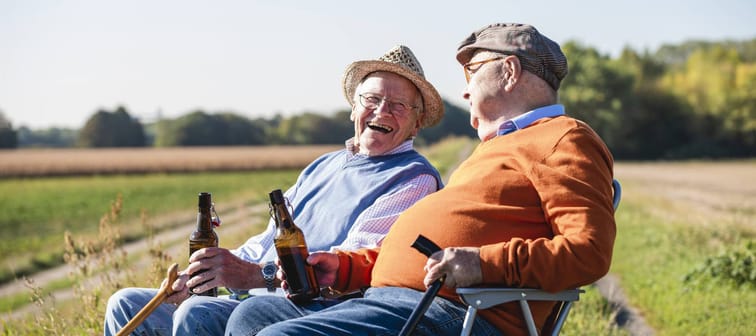 Two older men sit in a field and toast with drinks while smiling.