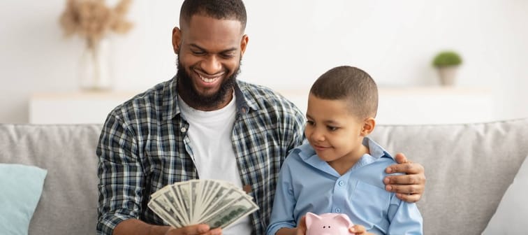 Father sitting with child on couch with fan of cash and piggy bank