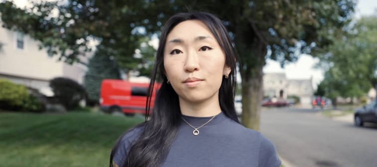 Woman with long black hair looks directly at camera, standing on sidewalk.