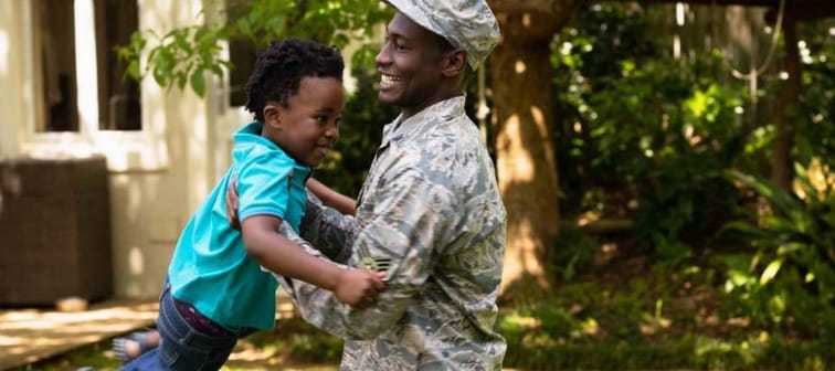 SIde view of a young adult African American male soldier in the garden outside his home, holding his young son and swinging him around, both smiling, their house in the background