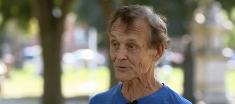 Man speaks directly to camera, standing outside at graveyard.