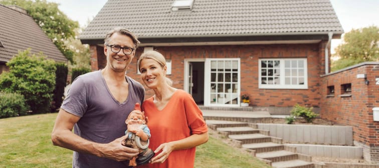 Happy couple poses on front lawn, holding a gnome in front of a brick house.