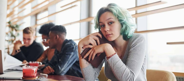 Young woman with green hair looks directly at camera, sitting at long table in coffee shop.