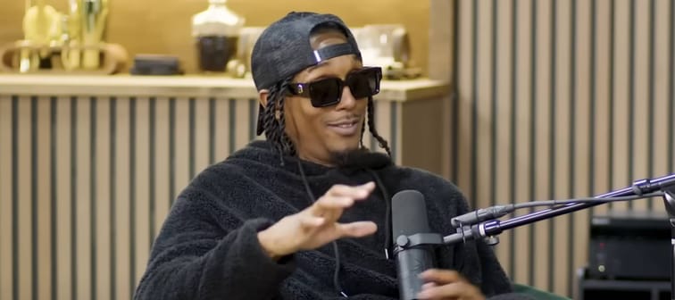 Former NBA star Lou Williams sits in chair on podcast set, wearing dark sunglasses and gesturing.