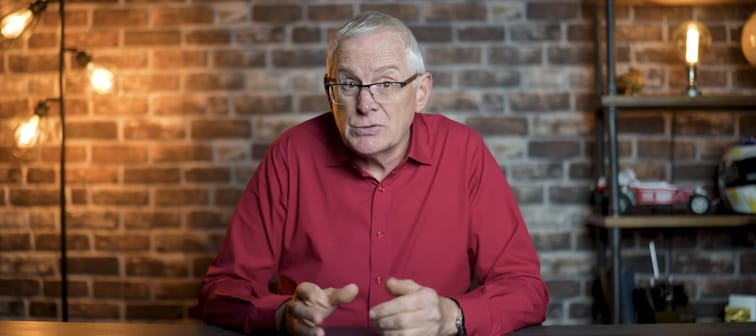 Man sits in front of brick wall, talking directly to the camera. Looking serious, intent.