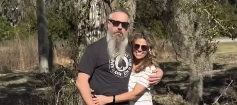 Couple pose with arms around each other in front of trees, outside.