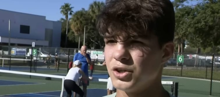 Young man stands on pickleball court, talking to camera, looking upset.