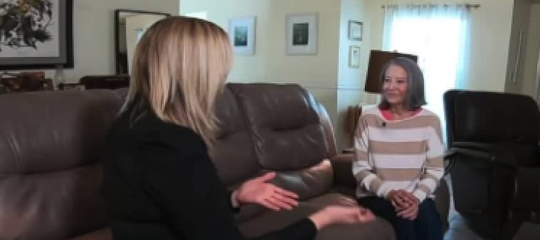 Two women sit facing each other on a couch in living room, talking.