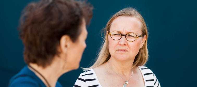 Two women seen facing each other, talking. One squinting at the other, looking skeptical.