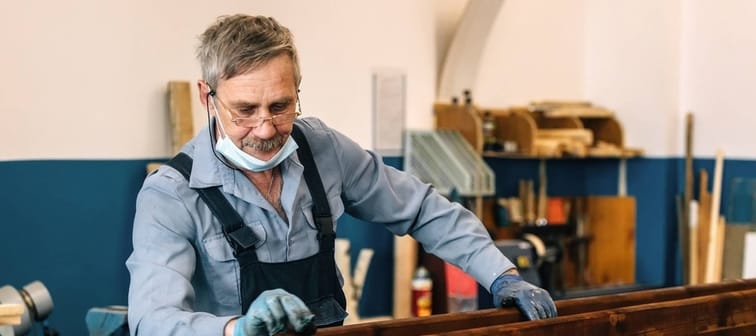 An elderly man paints wooden boards with dark varnish in a carpentry workshop.