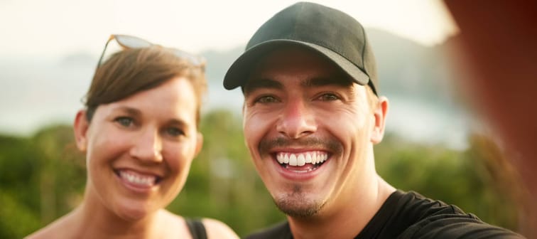 Young couple smile as they take a selfie outside.