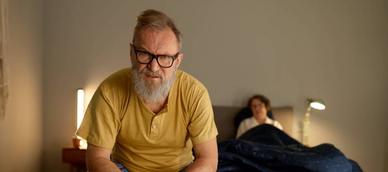 Man sits at the end of the bed with an incredulous look on his face, wife seen in the background sitting up in bed.