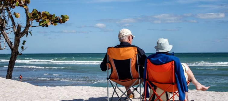 An older couple sit on folding chairs on a beach.