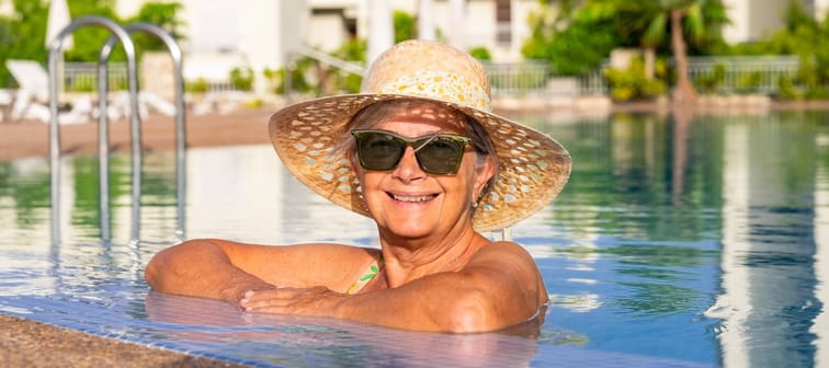 Older woman wearing straw hat and sunglasses smiles while leaning against side of pool.