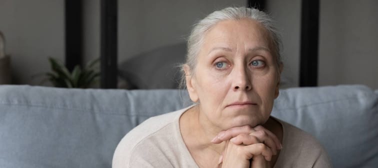 Sad lonely pensive elder lady looking away, sitting on couch at home with chin on hands