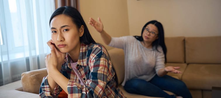 Two women fighting on the couch, one facing the camera looking upset, while another seen in the background looks angry.