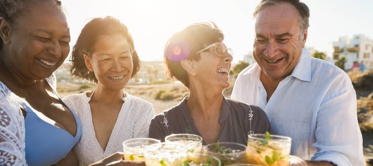 Senior multiracial friends cheering with alcohol free mojitos on the beach at sunset - Mature people having fun during summer vacation - Focus on right people faces