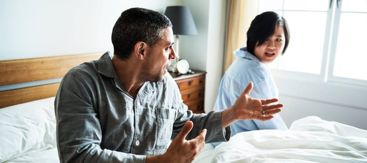 Couple in bed arguing, wearing pyjamas and gesturing angrily.