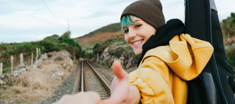 Woman wearing hat reaches out hand to the camera, leading forward.